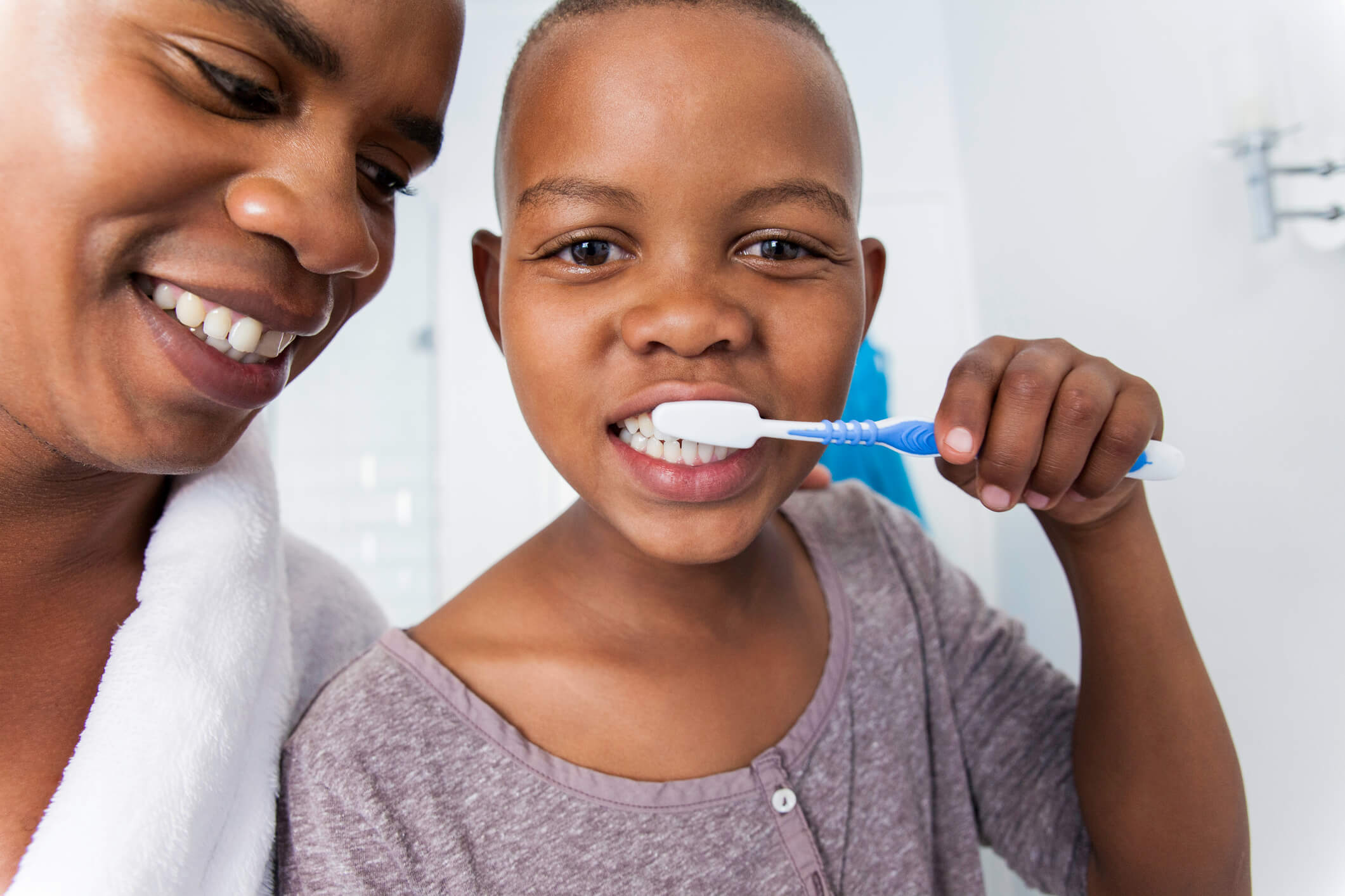 A parent smiles beside a child brushing their teeth at the bathroom sink